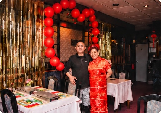 De eigenaren van Bao Long staan glimlachend in het restaurant, onder een boog van rode ballonnen.